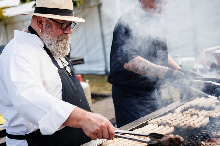 Koch mit Strohhut grillt Bratwürste und Steaks auf einem großen Grill bei einer Veranstaltung